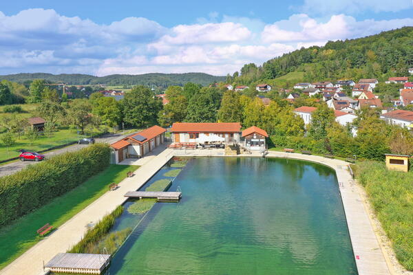Bild vergr&ouml;&szlig;ern: Auf dem Bild sieht man das Naturbad Königstein. Im Hintergrund sieht man den Ort Königstein und eine Toilette und mehrere Autos.