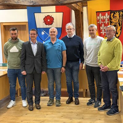Bild vergr&ouml;&szlig;ern: Bild mit 6 Erwachsenen vor zwei Wappen in einem Raum, welcher mit Holz verkleidet ist.
