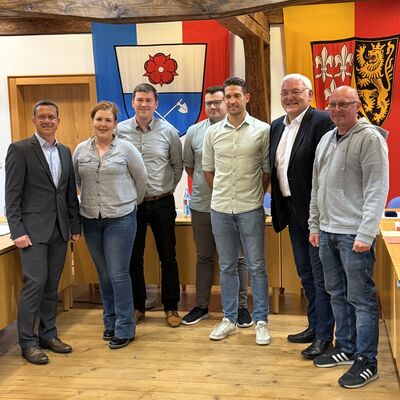Bild vergr&ouml;&szlig;ern: Bild mit 7 Erwachsenen vor zwei Wappen in einem Raum, welcher mit Holz verkleidet ist.
